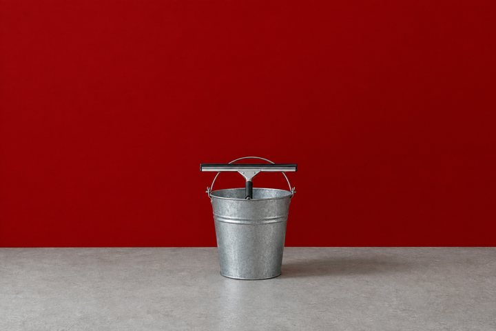 A metal bucket with a short window squeegee resting on its rim, on a grey concrete floor in front of a red wall.